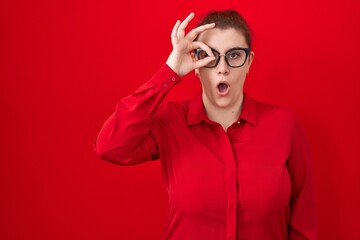 Young hispanic woman with red hair standing over red background doing ok gesture shocked with surprised face, eye looking through fingers. unbelieving expression.