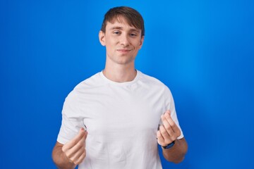 Caucasian blond man standing over blue background doing money gesture with hands, asking for salary payment, millionaire business