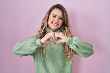 Young caucasian woman standing over pink background smiling in love doing heart symbol shape with hands. romantic concept.