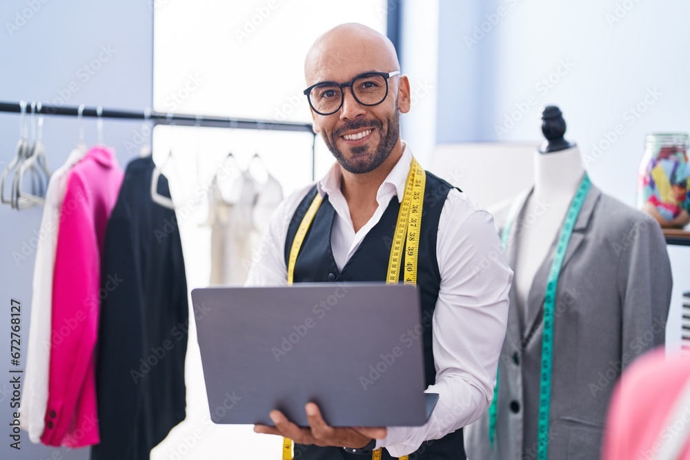 Poster young bald man tailor smiling confident using laptop at tailor shop