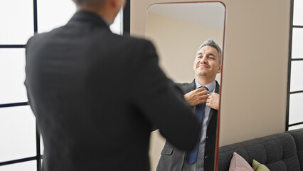 Young hispanic man business worker wearing suit looking on mirror at home