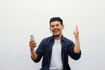 Young handsome Asian wearing casual shirt holding smartphone and pointing up with fingers while smiling confident and happy over white background