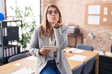Young hispanic woman working at the office wearing glasses looking at the camera blowing a kiss with hand on air being lovely and sexy. love expression.
