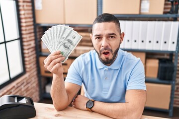 Hispanic man working at small business ecommerce holding dollars scared and amazed with open mouth for surprise, disbelief face