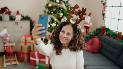 Middle age hispanic woman make selfie by smartphone sitting on floor by christmas tree at home