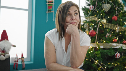 Middle age hispanic woman sitting on sofa by christmas tree looking upset at home