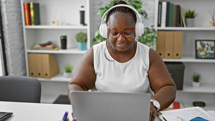 Brimming with success, this smiling african american boss-woman is slaying her business world. hunkered down in the office, jamming to music in headphones, she's working that laptop.