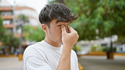 Exhausted young hispanic teenager rubbing itchy eyes amidst cool city park's green nature, a...