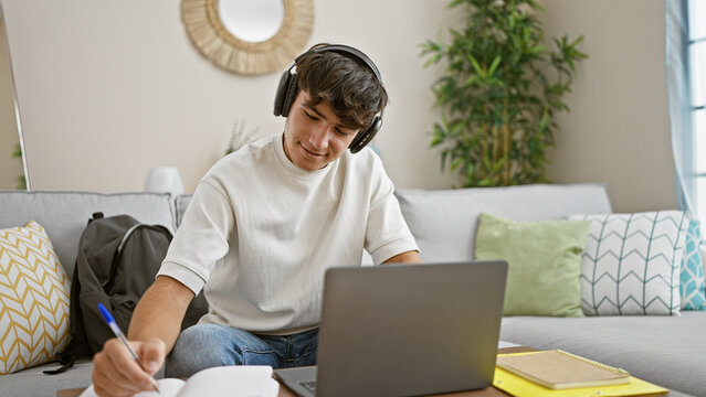 Confident young hispanic teenager lounging on living room sofa, passionately engaged in online education, using his laptop, diligently taking study notes at home
