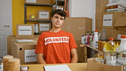 Young hispanic teenage volunteer with serious face sits at a table in charity center, showing true...