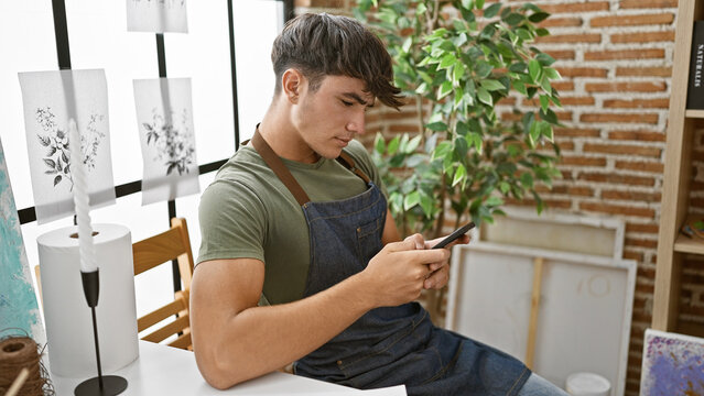Hispanic teen boy artist immersed in art lesson, sitting on studio chair, concentrating on painting and typing on smartphone