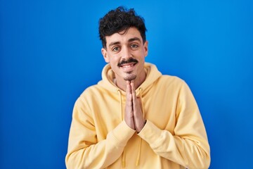 Hispanic man standing over blue background praying with hands together asking for forgiveness smiling confident.
