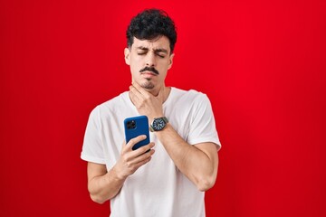 Hispanic man using smartphone over red background touching painful neck, sore throat for flu, clod and infection
