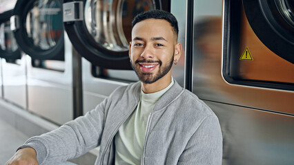 Young hispanic man smiling confident sitting on the floor at laundry facility - obrazy, fototapety, plakaty