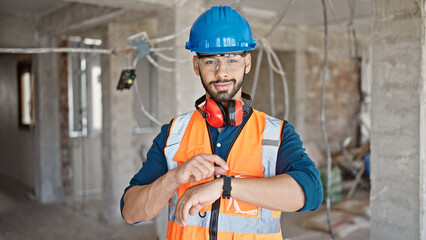 Young hispanic man builder looking at wrist watch at construction site