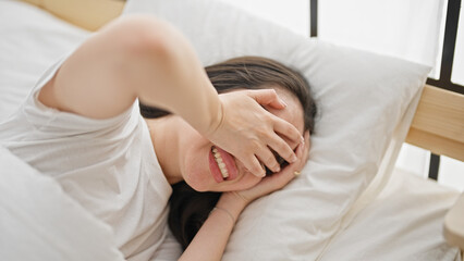 Young beautiful hispanic woman smiling confident covering eyes with hands lying on bed at bedroom