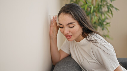Young beautiful hispanic woman listening through wall with glass at bedroom