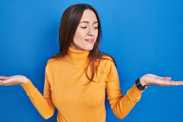 Young brunette woman standing over blue background smiling showing both hands open palms, presenting and advertising comparison and balance