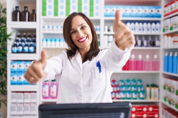 Middle age brunette woman working at pharmacy drugstore approving doing positive gesture with hand, thumbs up smiling and happy for success. winner gesture.