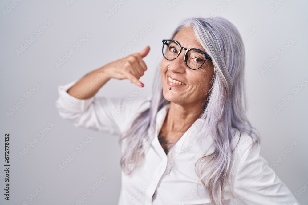 Poster middle age woman with tattoos wearing glasses standing over white background smiling doing phone ges