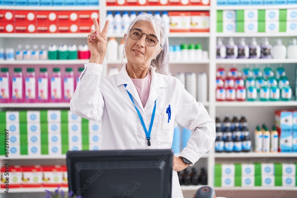 Sticker Middle age woman with tattoos working at pharmacy drugstore smiling looking to the camera showing fingers doing victory sign. number two.