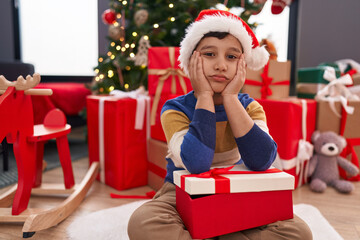 Adorable hispanic boy holding christmas gift sitting on floor with unhappy expression at home