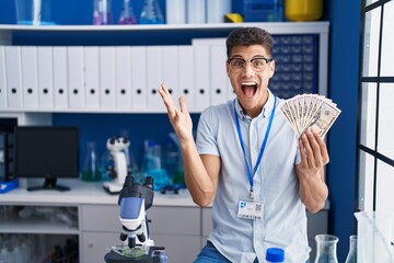 Naklejka na ściany i meble Young hispanic man working at scientist laboratory holding dollars celebrating victory with happy smile and winner expression with raised hands