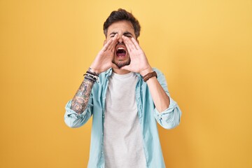 Young hispanic man with tattoos standing over yellow background shouting angry out loud with hands over mouth