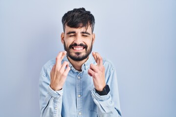Young hispanic man with beard standing over blue background gesturing finger crossed smiling with hope and eyes closed. luck and superstitious concept.