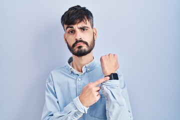 Young hispanic man with beard standing over blue background in hurry pointing to watch time,...