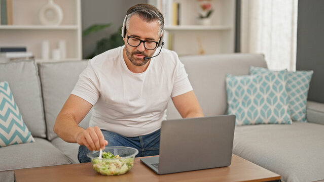 Grey-haired man eating salad using laptop and headphones at home