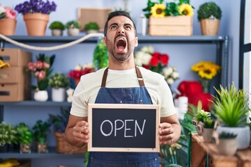 Hispanic man with beard working at florist holding open sign angry and mad screaming frustrated and furious, shouting with anger looking up.