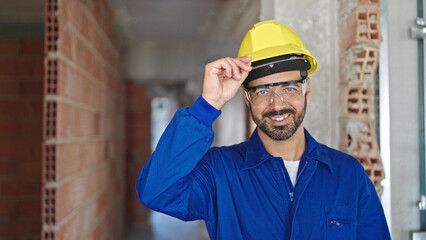 Young hispanic man worker wearing hardhat smiling at construction site