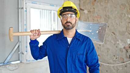 Young hispanic man worker wearing hardhat holding shovel at construction site