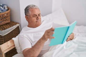 Middle age grey-haired man reading book sitting on bed at bedroom