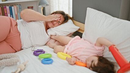 Obraz na płótnie Canvas Stressed mother and daughter lying in bed as baby plays with toys in bedroom - a poignant family moment