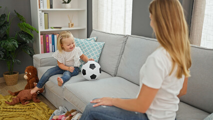 Happy caucasian mother and daughter playing ball together, sitting comfortably on living room sofa, indoors at home. smiling, relaxing family time.