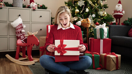 Obraz na płótnie Canvas Young blonde woman unpacking gift sitting on floor by christmas tree at home