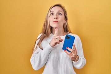Young blonde woman using smartphone typing message thinking concentrated about doubt with finger on...