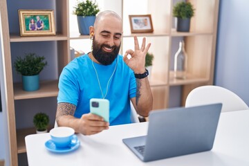 Young hispanic man with beard and tattoos doing video call with smartphone doing ok sign with fingers, smiling friendly gesturing excellent symbol