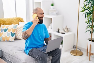 Young bald man talking on smartphone using laptop at home