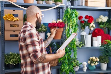 Young bald man florist reading document holding plant at flower shop