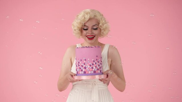 Attractive woman excitedly shows off birthday colorful cake, says wow. Woman celebrating birthday, victory. Woman in the image of surrounded by soap bubbles on a pink background.