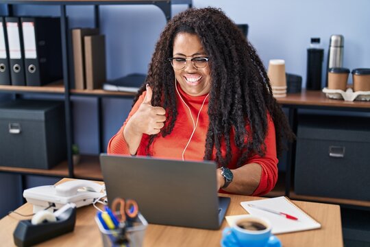 Plus size hispanic woman working at the office with headphones smiling happy and positive, thumb up doing excellent and approval sign
