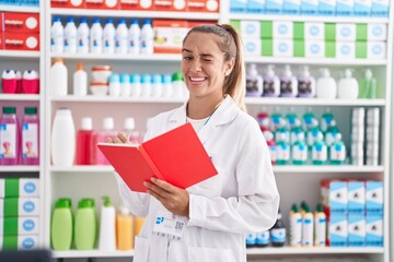 Young blonde woman working at pharmacy drugstore holding notebook winking looking at the camera...
