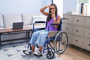 Young hispanic woman sitting on wheelchair at home smiling making frame with hands and fingers with...