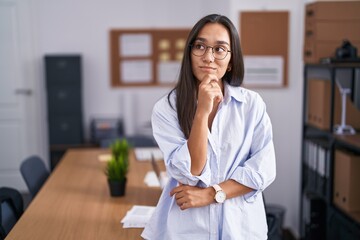 Young hispanic woman at the office with hand on chin thinking about question, pensive expression. smiling with thoughtful face. doubt concept. - obrazy, fototapety, plakaty