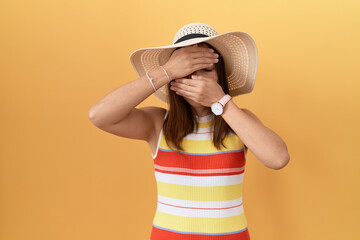 Middle age chinese woman wearing summer hat over yellow background covering eyes and mouth with hands, surprised and shocked. hiding emotion