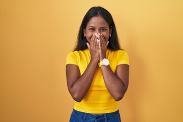 Young indian woman standing over yellow background laughing and embarrassed giggle covering mouth...