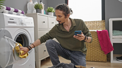 Young hispanic man using smartphone washing clothes at laundry room